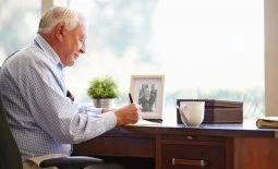 Senior Man Writing Memoirs In Book Sitting At Desk