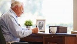 Senior Man Writing Memoirs In Book Sitting At Desk