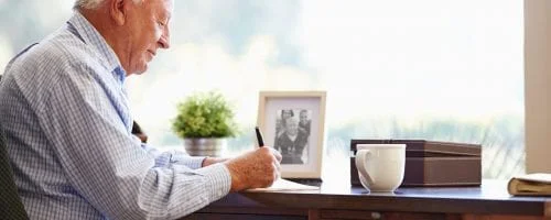 Senior Man Writing Memoirs In Book Sitting At Desk